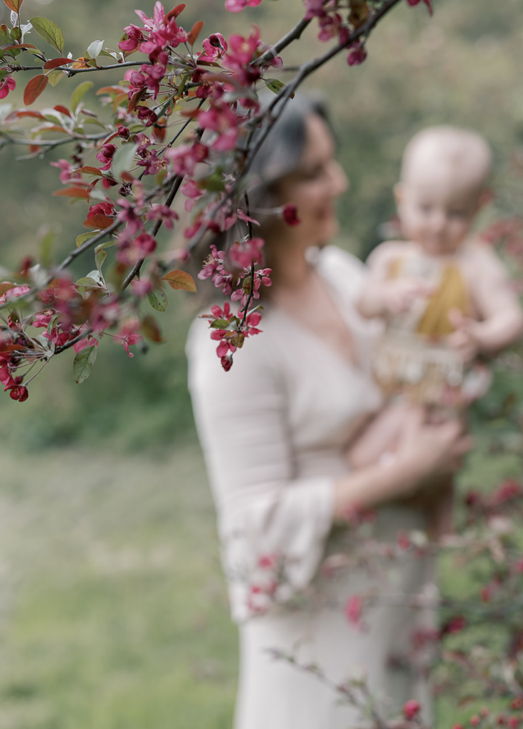 Flowering branch in foreground with mother and baby blurred in background during family photo session with Norabloom, Ithaca NY photographer